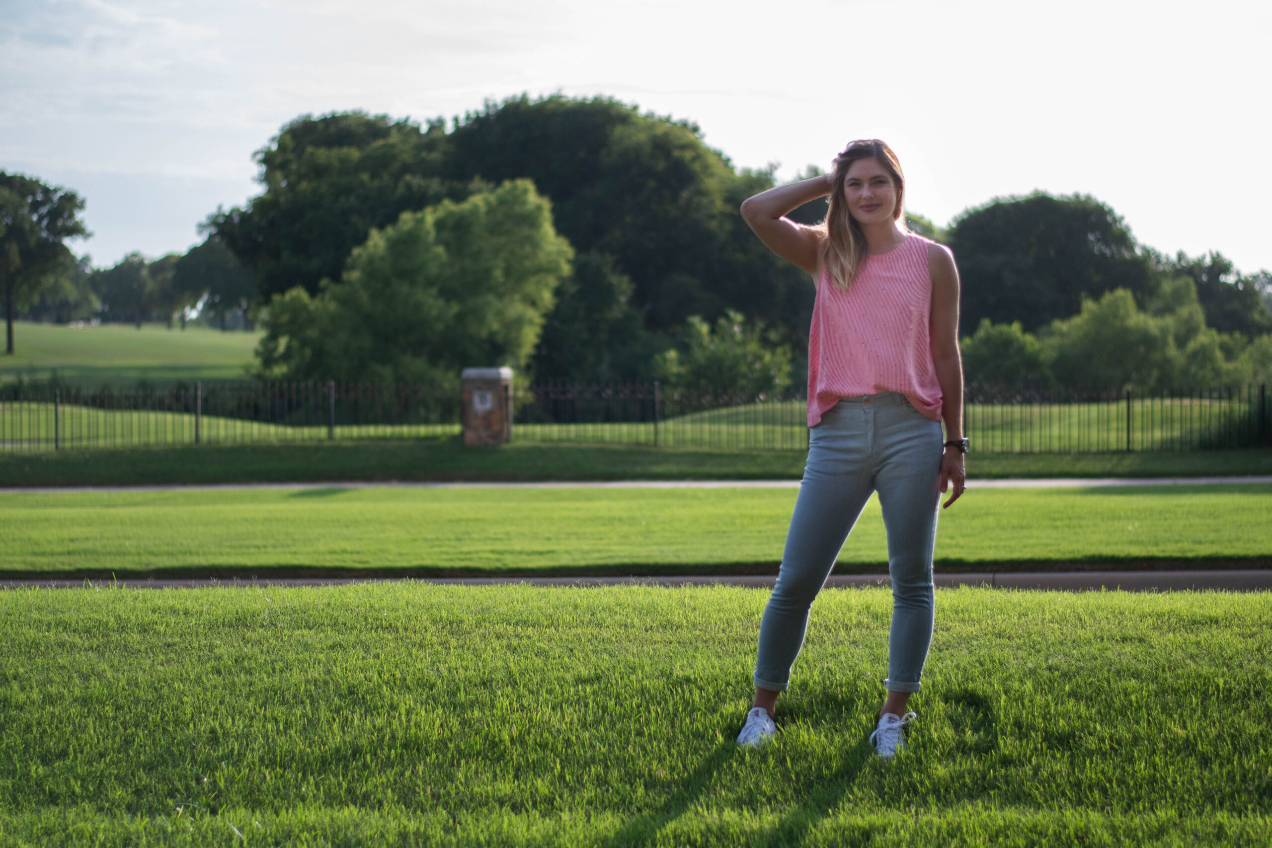Elizabeth stands on the grass during golden hour wearing a pair of light colored handmade denim jeans, a red and white striped tanktop, and white converse tennishoes. In the background are trees and bushes. Her stance leans slightly to her left, and her right arm is bent upwards, keeping her hair out of her face.