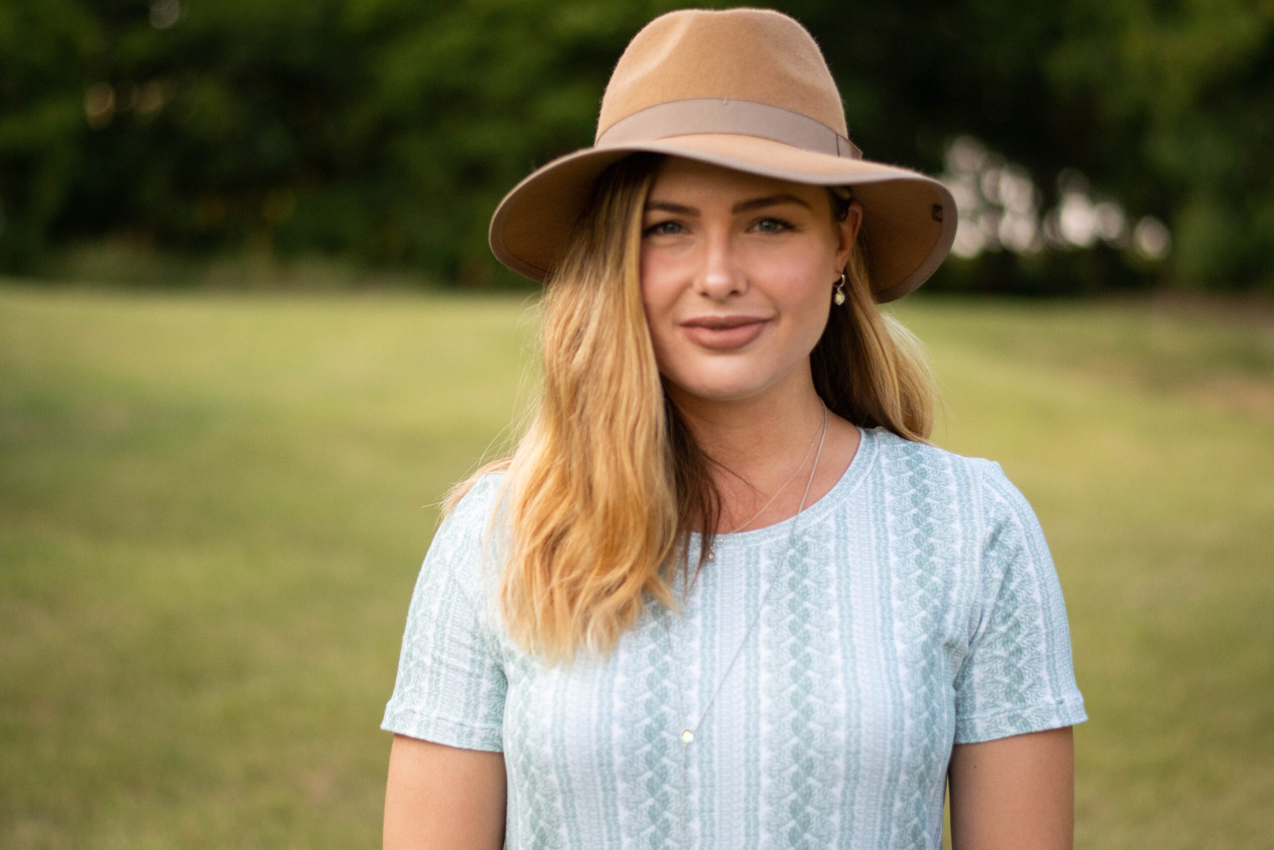 Elizabeth stands in front of a field of grass, wearing a camel colored hat and a t-shirt dress made out of a cable knit sweater look french terry fabric in a sage cream and cream color scheme